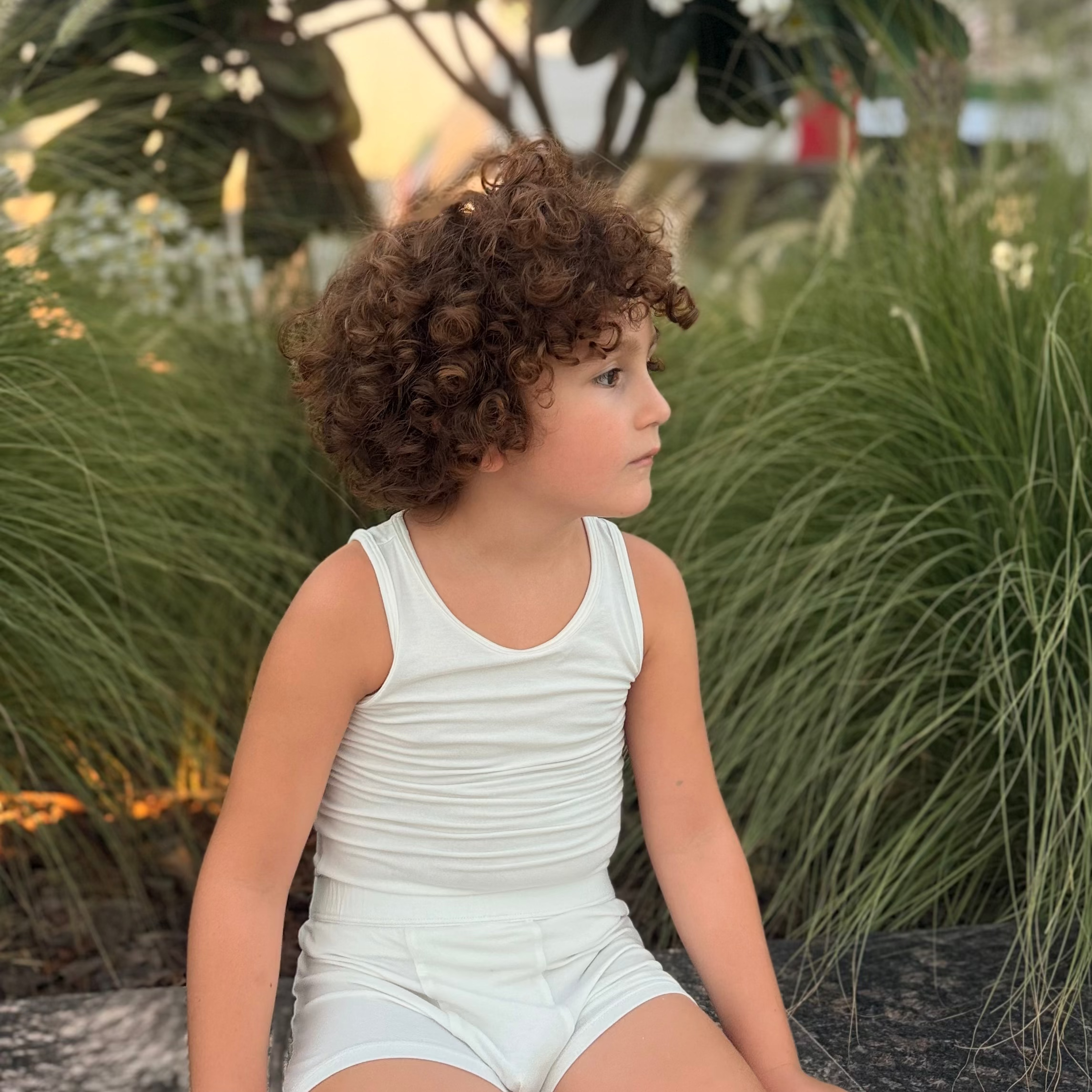 Child in a beige underwear sitting outdoors with greenery and flowers in the background