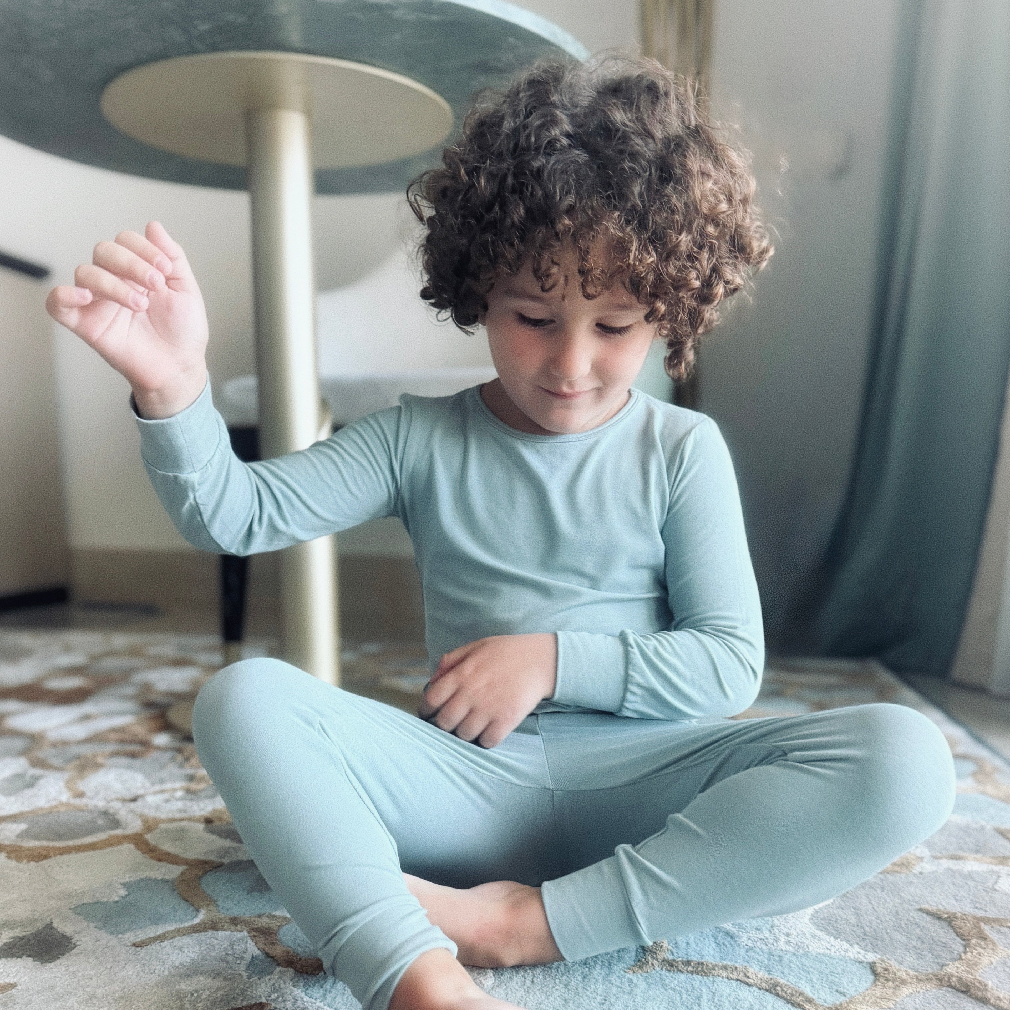 Child sitting on a patterned rug wearing light sage green pajamas.